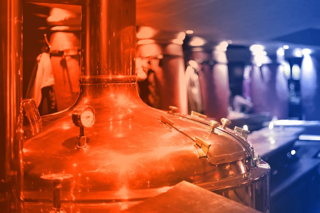 A brewery hall with large fermentation tanks. In the foreground is a detailed section of a fermentation tank with valves, flaps and a thermometer. Behind it are further tanks.