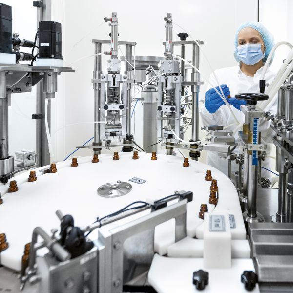 Darstellung einer mit ELKALUB H1-Schmierstoffen betriebenen Medizinabfüllanlage. Im Hintergrund arbeitet eine Technikerin in Schutzausrüstung.