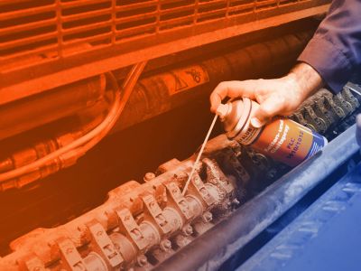 Close-up: View into the delivery of a printing machine. An employee sprays the joints of the delivery with a can of ELKALUB FLC 1010/1012 chain and sliding spray. Only the hand with the can is shown. A dosing aid is attached to the spray head.
