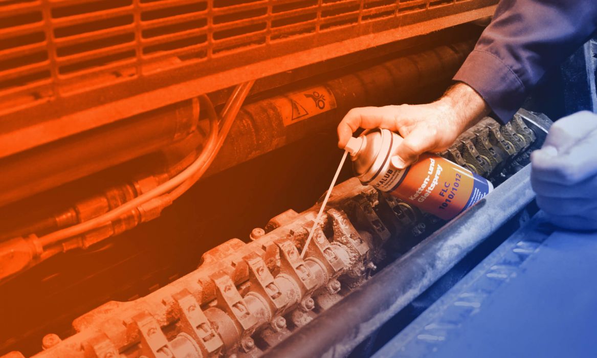 Close-up: View into the delivery of a printing machine. An employee sprays the joints of the delivery with a can of ELKALUB FLC 1010/1012 chain and sliding spray. Only the hand with the can is shown. A dosing aid is attached to the spray head.